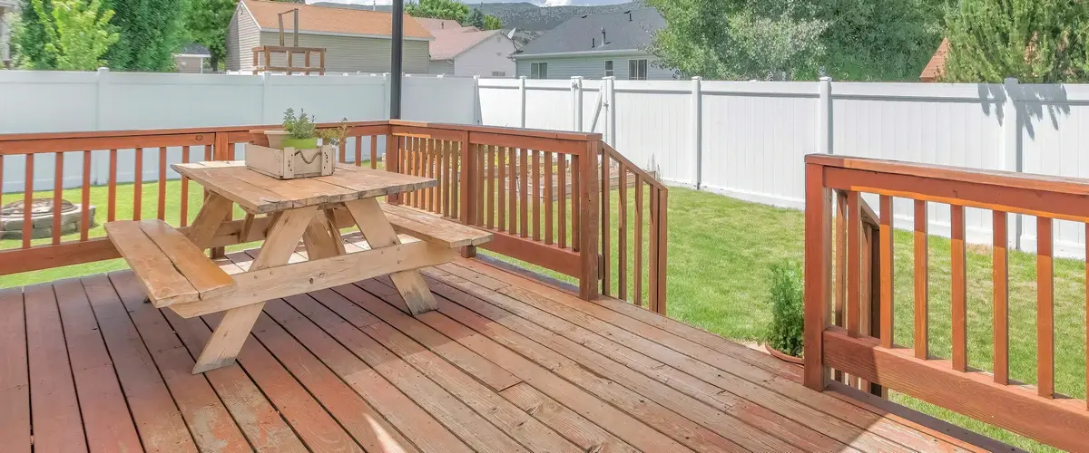 A spacious outdoor wooden deck featuring a rustic wooden picnic table, overlooking a green lawn and a white vinyl privacy fence.