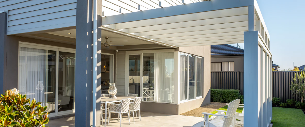 A contemporary home exterior featuring a white slatted covered patio structure over an outdoor dining area, looking out onto a manicured green lawn.
