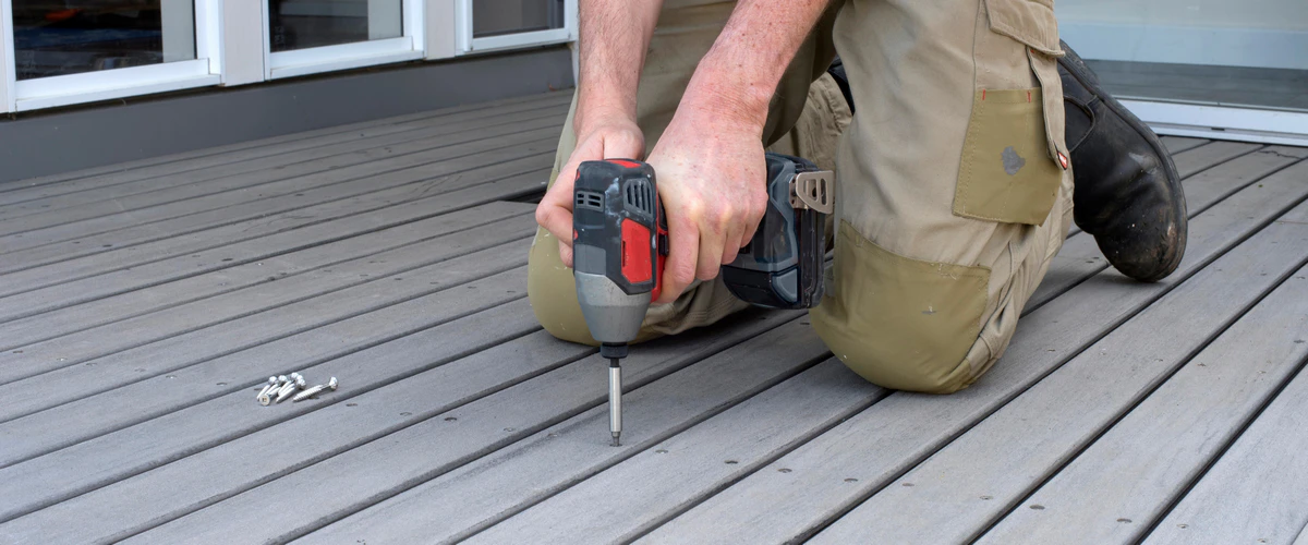 A contractor kneeling down during a deck repair, using an impact driver to securely fasten grey composite deck boards with a pile of stainless steel screws nearby.