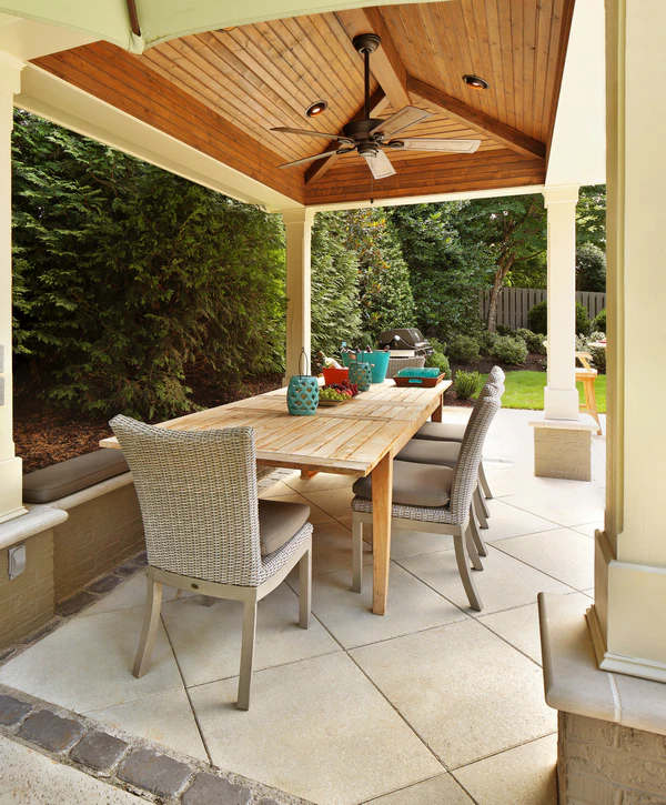 A spacious outdoor dining area built by a covered patio builder in Chattanooga, TN, featuring a beautiful vaulted wood ceiling, ceiling fan, and a long wooden table with wicker chairs.
