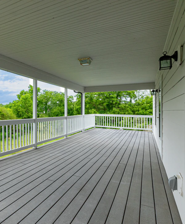 An expansive covered second-story composite deck with light grey flooring, white house siding, and a white ceiling with an antique central light fixture. Views of dense green trees. Built by local deck building companies in Chattanooga, TN.