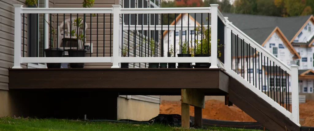 A low-elevation dark brown deck attached to a house featuring modern white railings with black metal balusters and a row of potted plants.