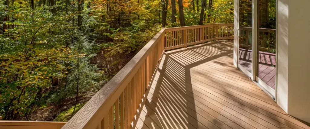 A spacious light brown wood deck connected to a house via sliding glass doors, casting long shadows while overlooking a lush, sunlit forest landscape.