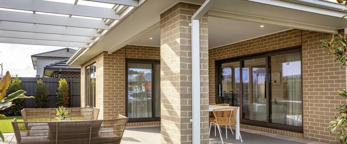 A modern brick house exterior showcasing a covered patio with a white slatted roof structure, featuring comfortable outdoor lounge seating and a dining set near glass sliding doors.