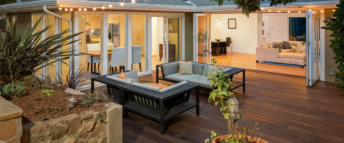 A modern outdoor deck at twilight illuminated by string lights, featuring dark patio lounge furniture and large open bi-fold doors leading into a brightly lit home interior.