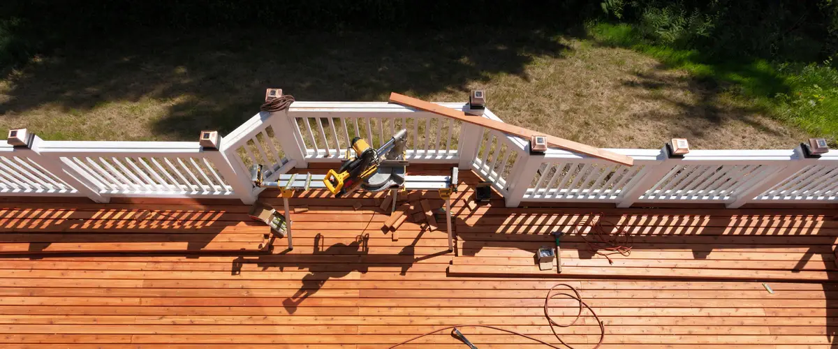 Aerial view of a residential wooden deck repair project in progress, showing freshly stained floorboards, white railings, and a miter saw cutting station.