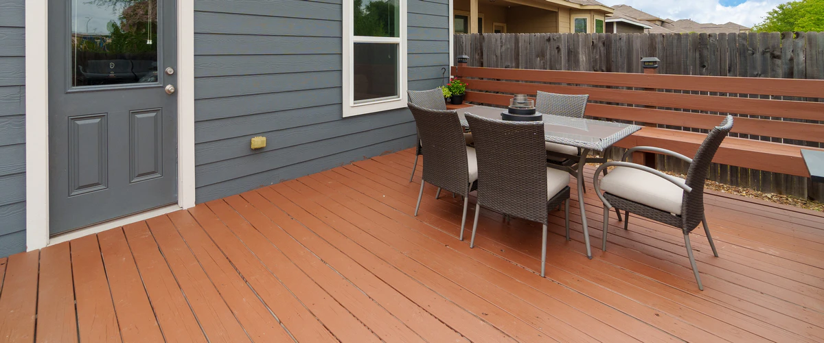 A painted reddish-brown outdoor deck attached to a grey house, furnished with a wicker dining set and featuring a built-in wooden bench.
