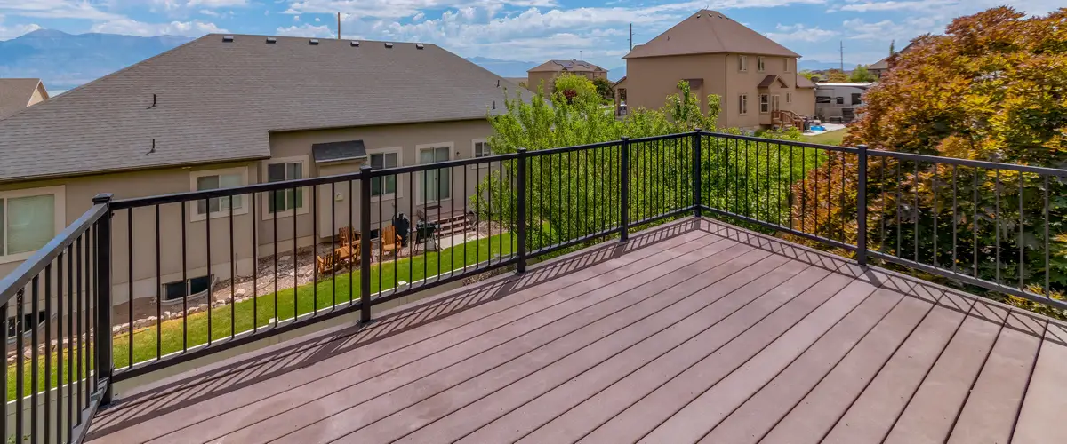 An empty, spacious composite deck featuring a sturdy black metal railing overlooking a grassy suburban backyard, neighboring homes, and distant mountains.