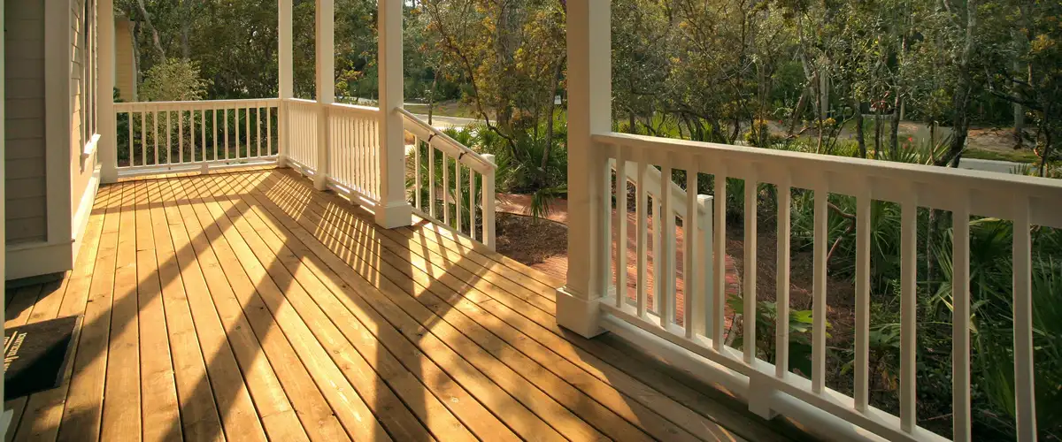 A traditional wooden deck and covered porch featuring white wooden railings, columns, and long shadows cast by the sun, looking out over a garden and brick pathway.