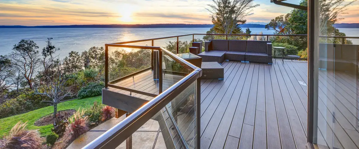 A stunning waterfront composite deck with a modern glass railing and comfortable brown wicker patio furniture, overlooking a large body of water at sunset.